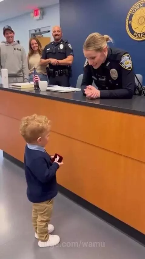Young boy in blue sweater proposing marriage to a female police officer at a police station counter.