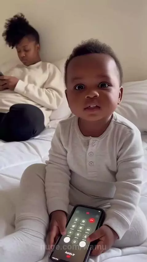 Baby in pajamas on a bed holding a smartphone, looking at the camera with wide eyes.