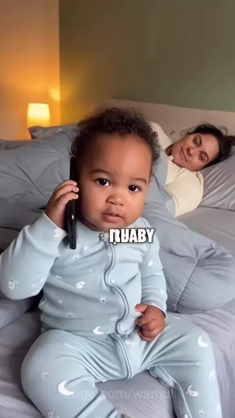 Baby with curly hair in a blue onesie holds a phone to its ear, talking to daddy while mom laughs in the background.