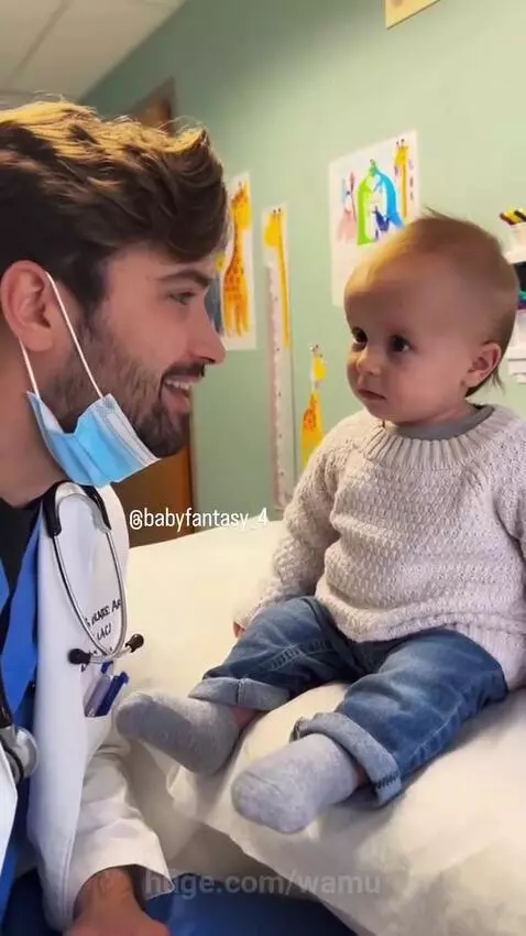 Doctor wearing a mask and white coat playfully interacting with a baby on an examination table, both laughing.