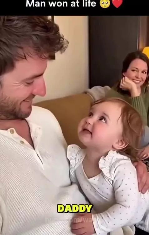 A baby girl sits on her father's lap, both smiling, while she playfully claims him as her own.