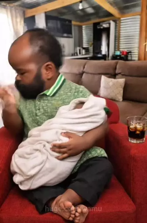 Man with dwarfism in a green polo shirt sitting in a red armchair, making funny faces at a swaddled baby.