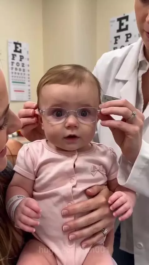 Baby smiling brightly while wearing new glasses, with an eye chart in the background.