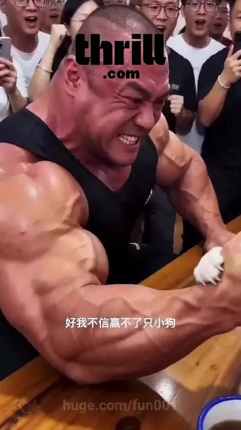 Muscular man arm wrestling a small fluffy dog wearing a bandana, with a cheering crowd.