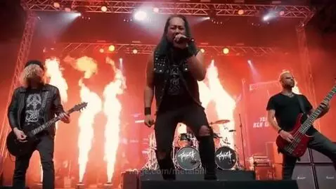 Heavy metal band performing live on stage with dramatic pyrotechnics, singer center, guitarist left, bassist right.