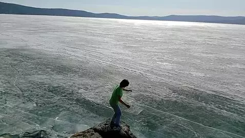 Person in green shirt and jeans on cliff edge, preparing to jump into a frozen lake with visible ice cracks.