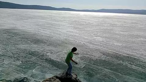 Woman in green t-shirt and jeans standing on a rocky cliff overlooking a frozen lake, gesturing towards the ice.