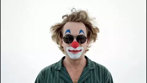 A clown maniacally presses a red button while a man in a suit looks on in distress, with surreal reality shifts in the background.