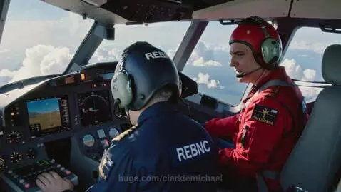 Two pilots in a modern aircraft cockpit, one in a blue uniform, the other in a red flight suit, flying above clouds.