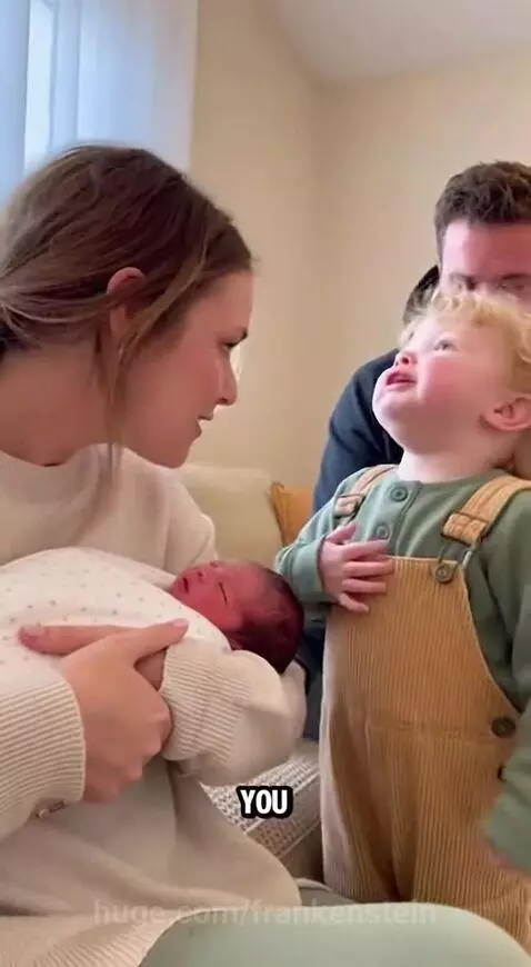 Mother holds newborn baby, toddler son looks on jealously, then kisses baby.