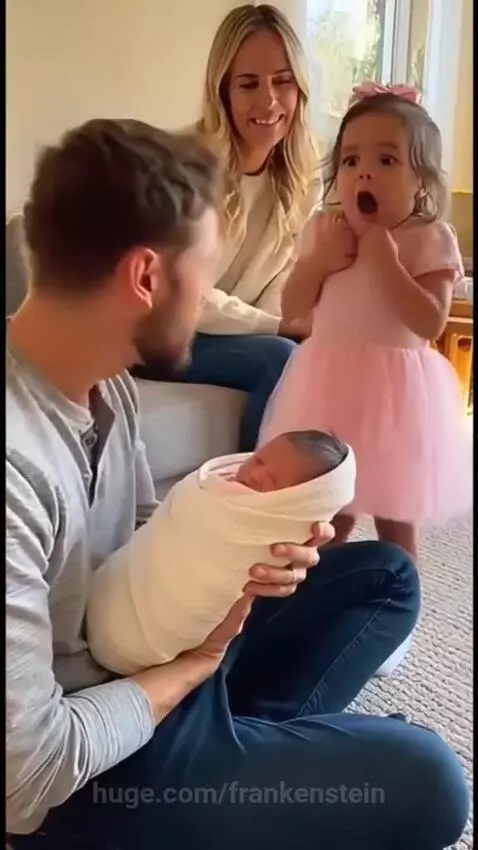 Toddler girl with shocked expression looks at a newborn baby held by her father.