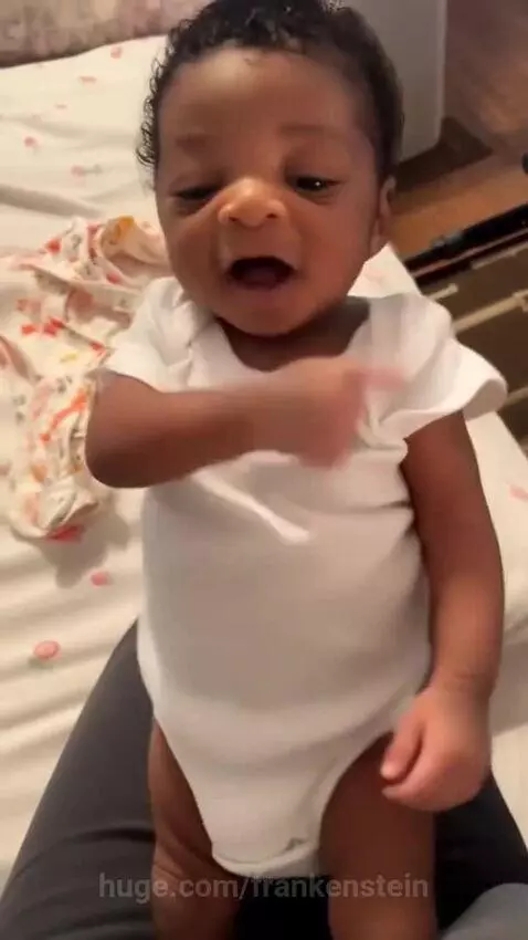 An infant with dark curly hair in a white onesie smiles and gestures while sitting on a lap, appearing to dance to music.