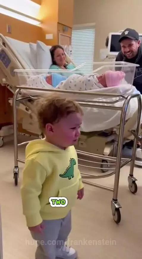 Toddler in yellow dinosaur hoodie crying in hospital, looking at parents with new bassinets.