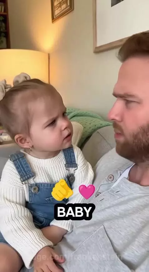 Young girl in overalls and sweater asks a bearded man in a grey shirt if he is 'mommy's baby'.