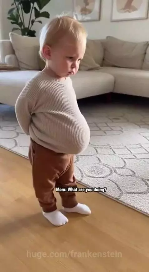 Blonde toddler in oversized sweater and brown pants stands on wooden floor, looking like a little grandpa.