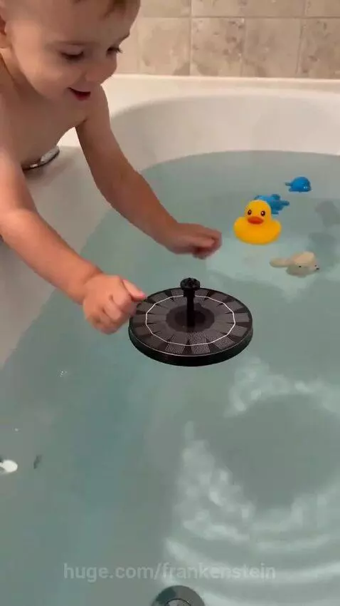 A smiling toddler in a towel watches a colorful, spraying light-up fountain bath toy.