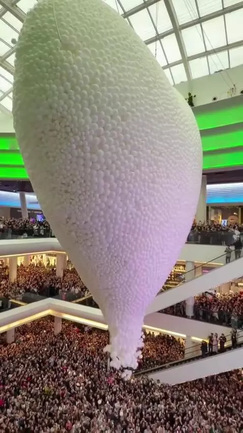 Thousands of white balloons falling from a net in a multi-level shopping mall, with excited crowds watching.