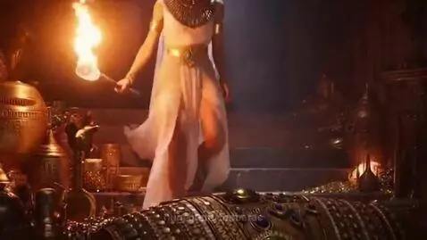 Woman in ancient Egyptian costume holding a torch, walking down steps in a treasure-filled chamber.