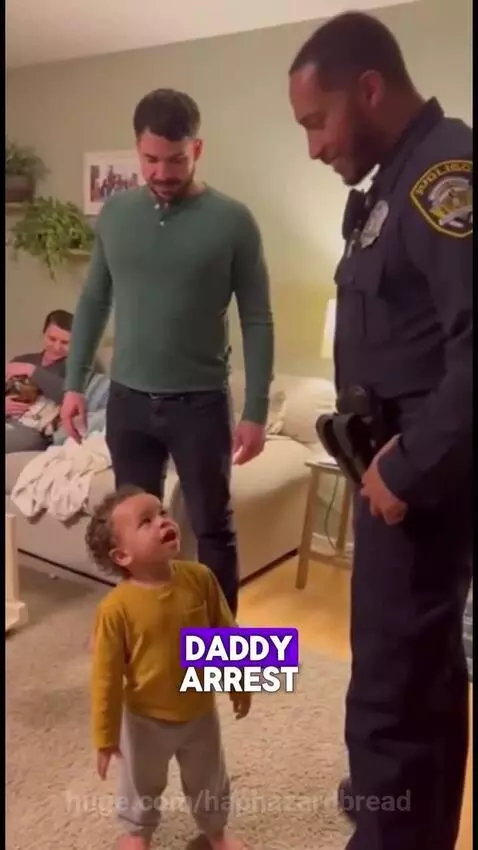 A young child talks to a police officer, with their dad behind them, in a living room.