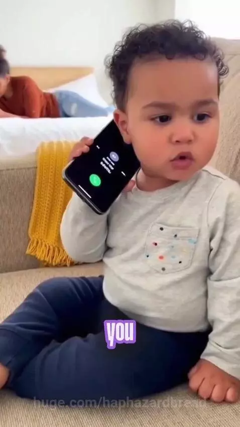 Baby with curly hair holding a smartphone to his ear, humorously complaining about diapers.