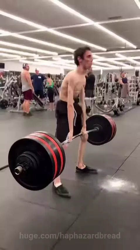 A very thin man in black shorts successfully performs a heavy deadlift in a gym, surprising onlookers.