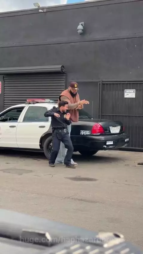 Police officer struggling to handcuff a much larger man next to a police car.
