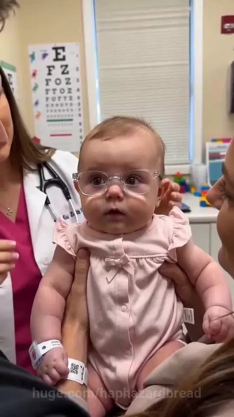 Baby wearing glasses smiles joyfully at parents for the first time.