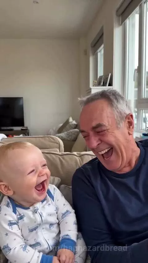 Baby and grandpa sitting on a couch, both laughing heartily at each other in a bright living room.