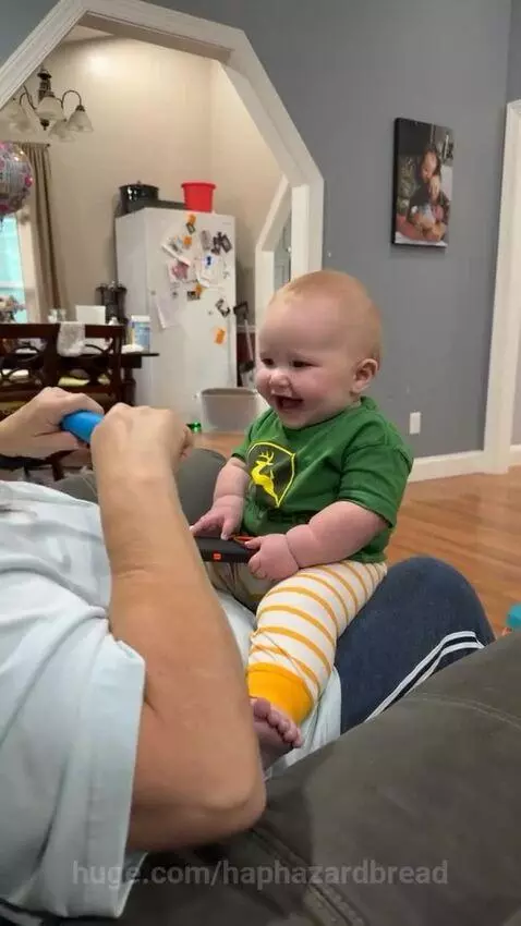 A baby wearing a green John Deere shirt laughs heartily while sitting on an adult's lap during a counting game.