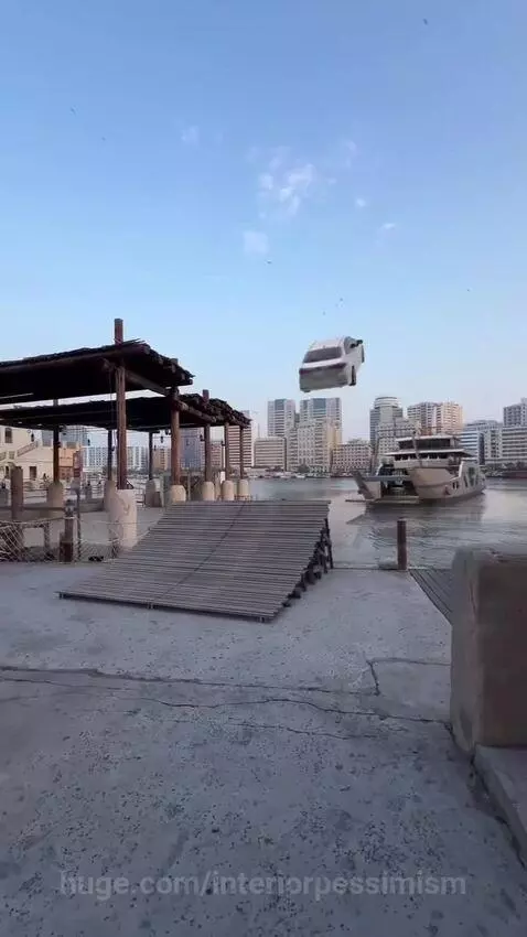 A white car flying through the air over water, launched from a ramp with city buildings in the background.