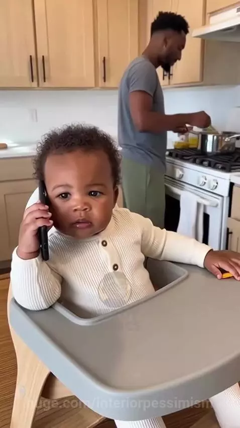 Baby in a high chair holding a smartphone to its ear, asking mom to come home because dad can't cook.