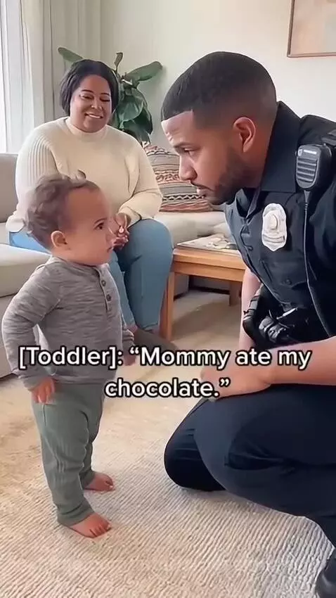 Toddler points at woman while police officer kneels to talk, woman laughs in background.