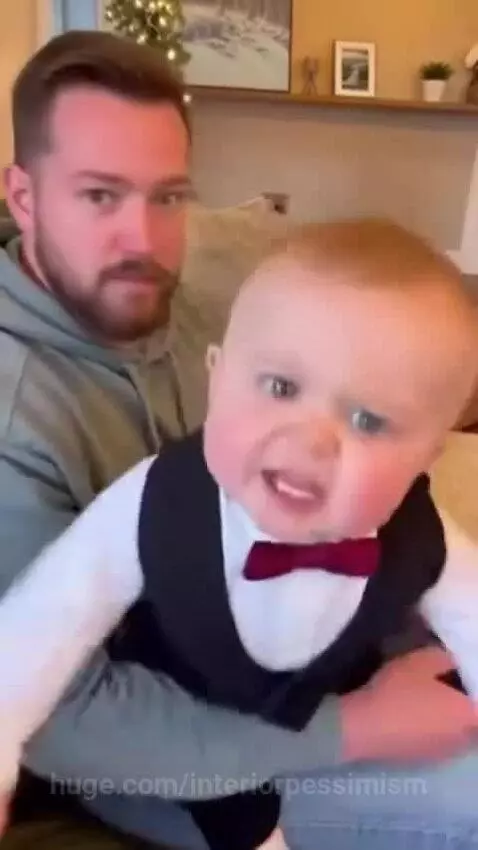 Baby in a bow tie glares angrily while sitting on a couch, with a woman's hand pointing and a man smiling in the background.