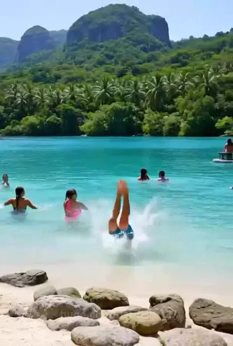 People enjoying a sunny day at a tropical beach with clear turquoise water and mountains. A man dives into the water.