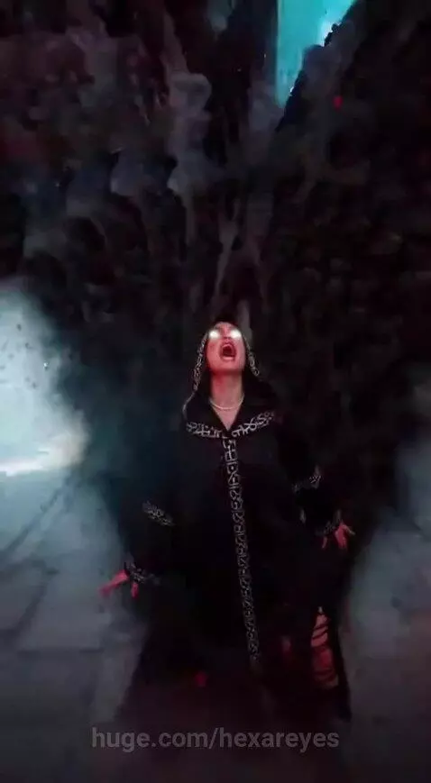 Person in a hooded robe with silver patterns performs magic with glowing white eyes and red lightning in a gothic hall.