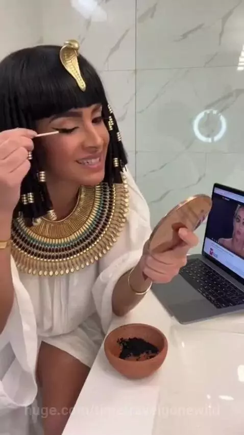 Woman dressed as Cleopatra attempts to apply eyeliner using a tutorial, holding a mirror and brush.
