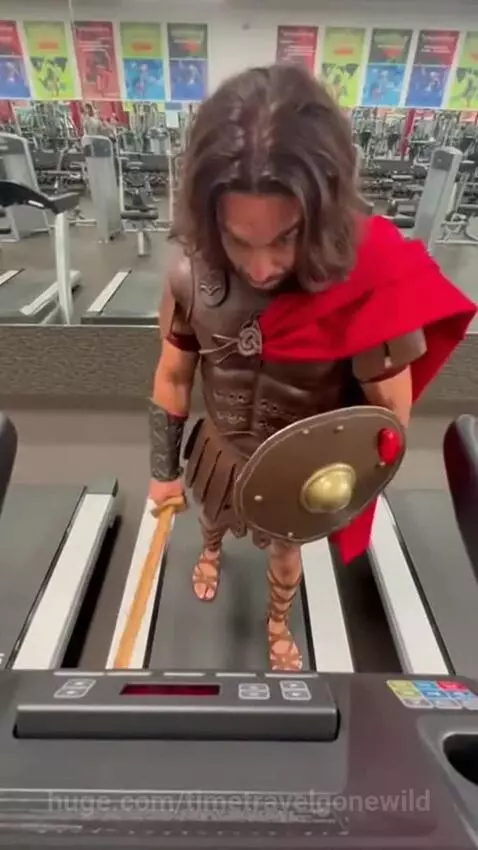 Man dressed as a Roman gladiator on a treadmill in a gym, holding a wooden sword and shield.
