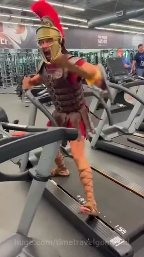 Man dressed as a Roman gladiator running on a treadmill in a gym, wearing armor and helmet.
