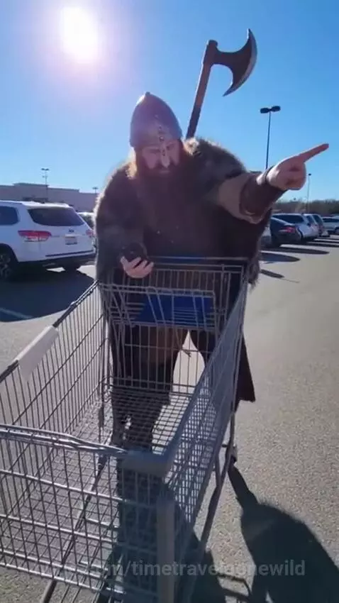 Man dressed as a Viking in a shopping cart looks at his phone, using GPS navigation in a sunny parking lot.