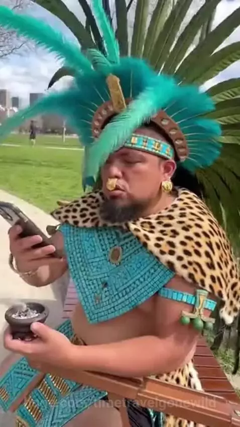 Aztec priest in feathered headdress holding a smartphone and censer, looking distressed at the weather app.