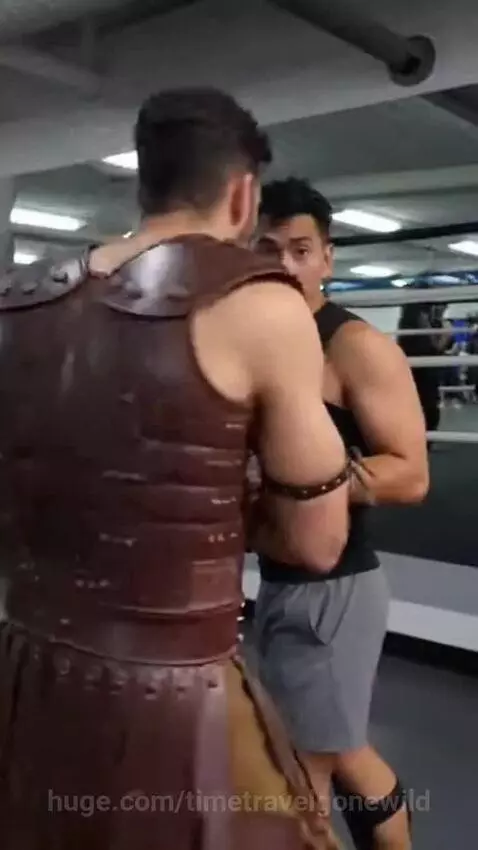 Man in gladiator costume with beard in a gym, observing other men working out.