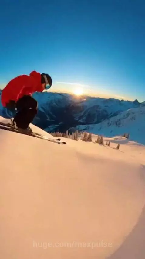 Skier in red jacket performs a jump off a snow mound on a mountain slope at sunset, with powder spraying.