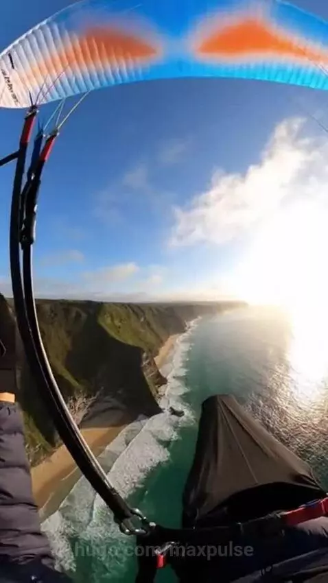 First-person view of paragliding over a scenic coastline with colorful canopy, turquoise ocean, and green cliffs.