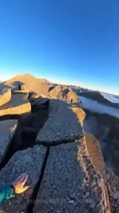 First-person view of a person reaching a mountain summit, revealing a panoramic view of mountains and clouds.