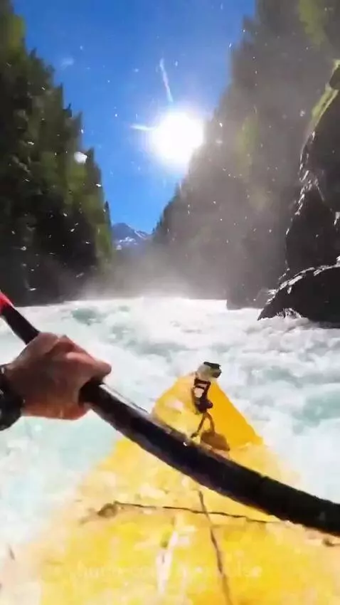 First-person view of a yellow kayak navigating turbulent whitewater rapids with mountains in the background.