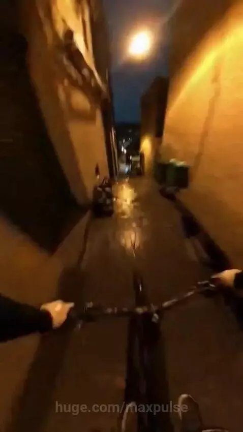 First-person view of cycling down a narrow, wet, dimly lit alley at night with streetlamp reflections.