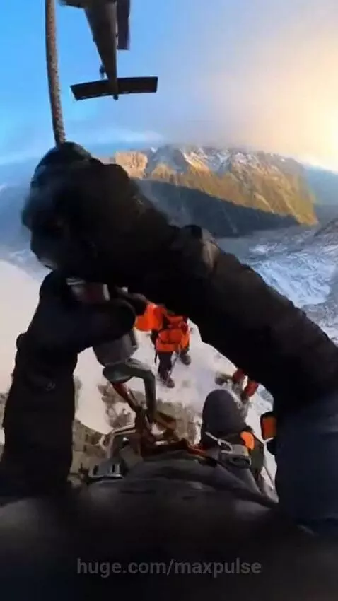 First-person view of a helicopter rescue hoist from a snowy mountain peak with clouds and blue sky.