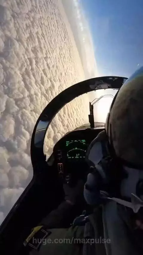 First-person view from a fighter jet cockpit flying high above white clouds, with the sun visible.
