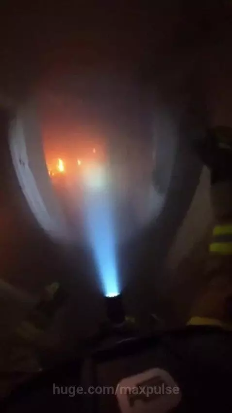 Firefighter's helmet camera view inside a dark, smoke-filled burning building with visible flames and a thermal imaging beam.