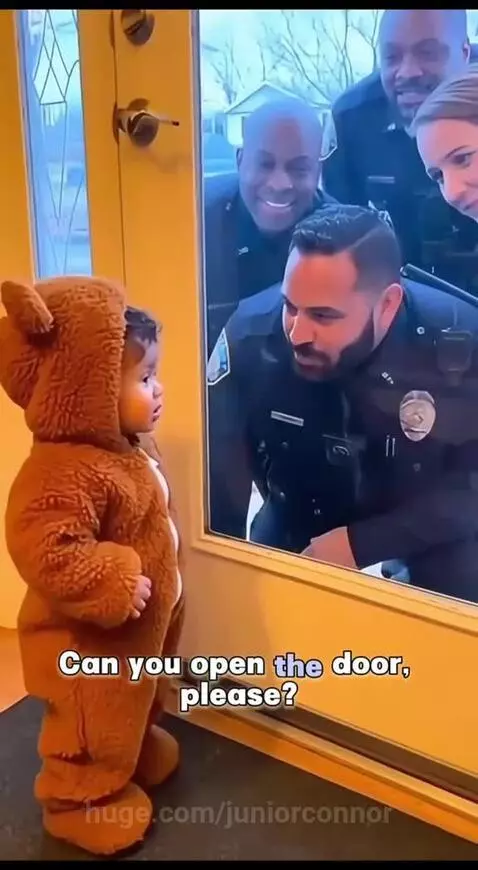 Child in a brown bear costume inside a house, looking through a glass door at smiling police officers.